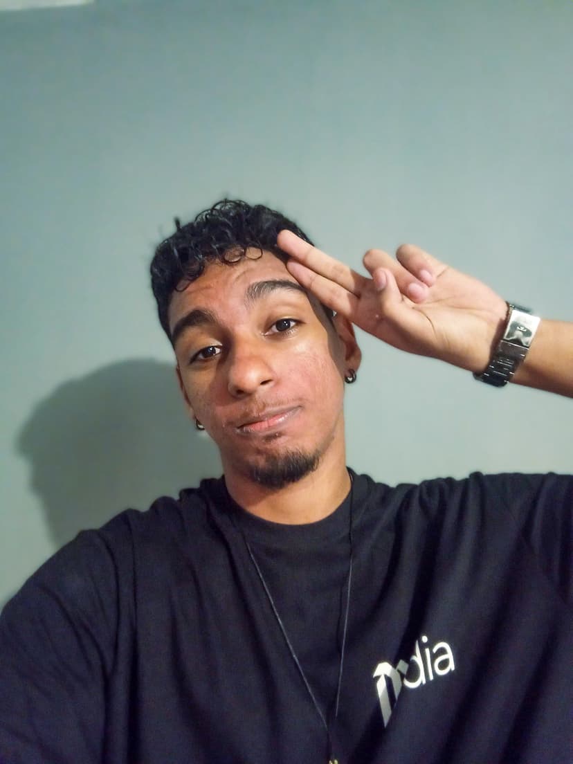Eu, jovem de pele parda e cabelos cacheados preto curtos, usando uma camiseta preta, com um leve sorriso no rosto, fazendo sinal com a mão ✌.